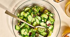 a glass bowl filled with sliced cucumbers next to two cups of tea and spoons