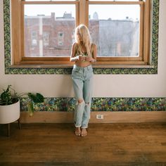 a woman standing in front of a window looking at her cell phone while wearing ripped jeans