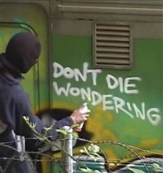 a man in a black hoodie writing on the side of a green wall with barbed wire