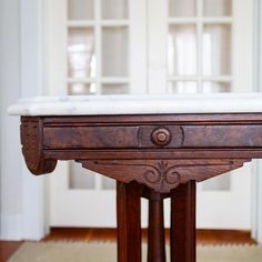 an old wooden table with marble top in front of a white wall and wood floors