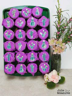 purple cupcakes with cactus designs on them sitting in a green tray next to flowers