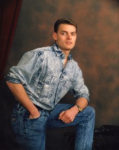 a young man sitting on top of a chair wearing jeans and a denim shirt with his hands in his pockets