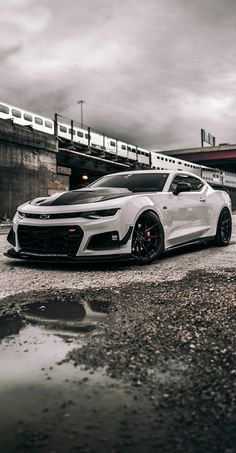 a white chevrolet camaro parked in front of a train on a track near a bridge