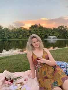 a woman sitting on the grass next to a lake holding a wine glass in her hand