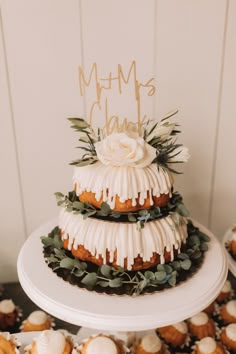 a three tiered cake with white icing and greenery on top, surrounded by cupcakes