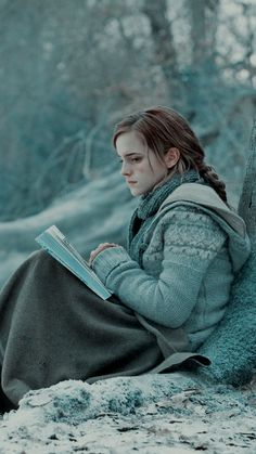 a young woman sitting on the ground reading a book in winter time, with trees and snow behind her