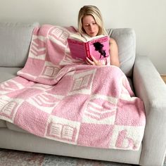 a woman sitting on a couch while reading a book under a pink and white blanket