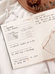 an open notebook sitting on top of a white bed next to glasses and flowers in a vase