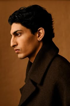 a man with black hair wearing a pea coat and looking off to the side in front of a brown background