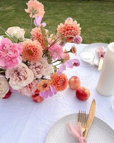 an arrangement of flowers and fruit on a white table cloth with pink napkins, goldware