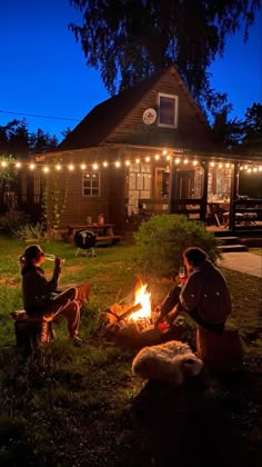 two people sitting around a campfire with lights strung over the house in the background