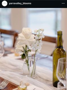 the table is set with white flowers in vases and wine bottles on it, along with silverware