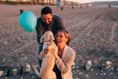 a man and woman holding a dog on the beach with balloons in front of them