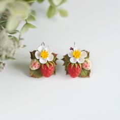 two small earrings with flowers on them sitting next to some leaves and berries in front of a white background