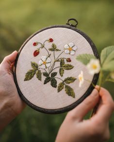 a person holding up a cross - stitched embroidery project with white flowers and green leaves