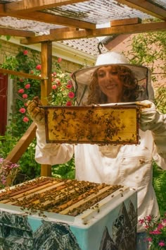 a woman in bee suit holding up a frame with bees on it and flowers around her