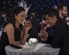 a man and woman sitting at a table with drinks in front of them, talking to each other