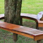 two wooden benches sitting next to each other in the grass near a tree with a hat on it