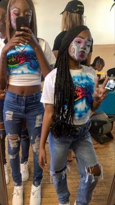 two girls with face paint standing in front of a mirror looking at their cell phones