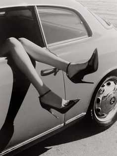 a black and white photo of a woman in high heels leaning on the door of a car