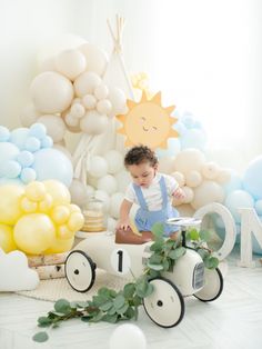 a baby boy sitting in a toy car surrounded by balloons and greenery on the floor