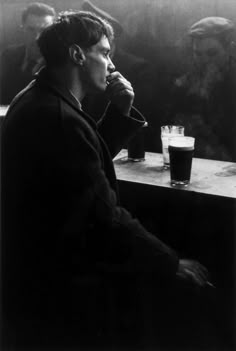 a black and white photo of a man sitting at a table with a drink in front of him