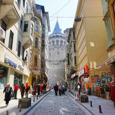 people are walking down an old cobblestone street with buildings on both sides and a tower in the background