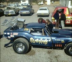 an old race car is parked in a parking lot with other cars and people around it