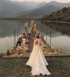 a woman in a wedding dress is standing on the edge of a dock with mountains in the background