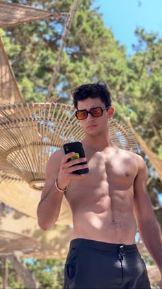 a shirtless man holding a cell phone in his right hand and wearing swim trunks