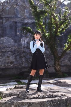 a girl in a short black dress and knee high boots standing on rocks with her arms crossed