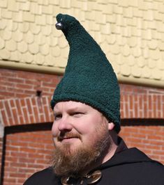 a bearded man wearing a green hat with a bird on it's head, standing in front of a brick building