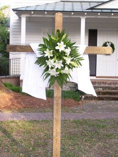 a cross decorated with white flowers and greenery in front of a house on a sunny day