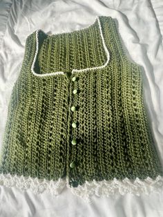 a green knitted vest laying on top of a white bed spread with buttons and lace