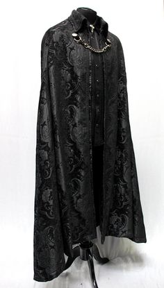 "A magnificent cape fit for royalty. Made in rich flowing black tapestry fabric with rich black satin lining inside. The high pointed collar can be worn down or up for an extra dramatic effect. Braided black trim lines the collar and along the front of cape. Fully lined in rich black satin. Fastens in front with a heavy antiqued chain which clasp at either side to heavy D-rings and decorated with two ornate metal buttons. Slits in the front of cape allow the wearer to reach through and hold a be Dark Robes Male, Evil King Outfit, Masquerade Ball Outfit Men, Wedding Cape Men, Mens Vampire Fashion, Elegant Fitted Cape For Costume Party, Formal Black Cape With Cape Sleeves, Elegant Black Cape For Costume, Formal Fitted Black Cape