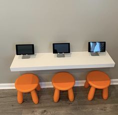 three orange stools sitting next to each other in front of a white table with two monitors on it