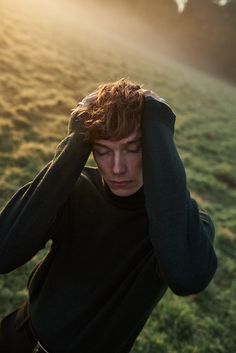 a young man with his hands on his head in the grass, looking off to the side