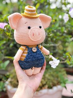 a hand holding a small crocheted pig toy in front of some potted plants