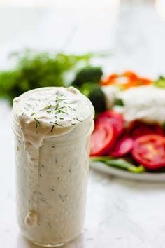 a jar filled with white sauce sitting on top of a table next to a plate of vegetables