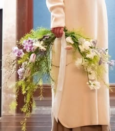 a woman wearing a white coat holding flowers and greenery in her hand while standing on a wooden floor