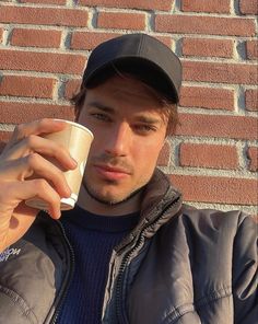 a man holding a coffee cup in front of a brick wall and wearing a black hat