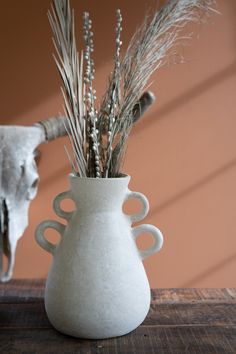 a white vase filled with dried plants on top of a wooden table next to a cow skull