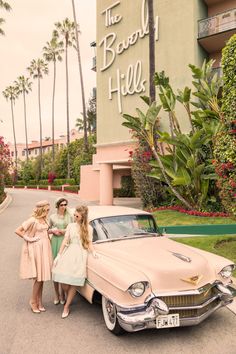 three women standing next to an old car in front of a hotel