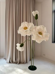 three white flowers are in a vase on the floor next to a curtained window