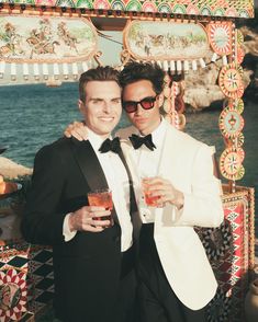 two men in tuxedos pose for a photo at an outdoor event by the water