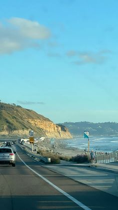 cars are driving down the road by the beach and cliffs in the distance with people standing on the sidewalk looking out at the water