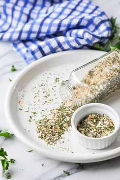 a white plate topped with a jar filled with seasoning next to a bowl of herbs