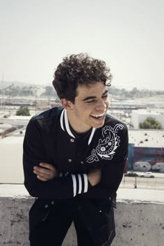 a man with curly hair is smiling and leaning on a wall in front of the city