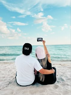 a man and woman sitting on top of a sandy beach next to the ocean holding up a camera