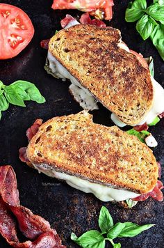 two toasted sandwiches with bacon, cheese and basil on a black surface next to tomatoes